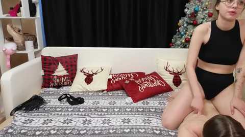 Media: Video of a slender, fair-skinned woman with glasses wearing a black crop top and panties, sitting on a man's face on a bed decorated with red and white holiday pillows.