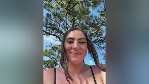 Media: Video of a young woman with long brown hair, wearing a black tank top, smiling outdoors with a clear blue sky and pine trees in the background.