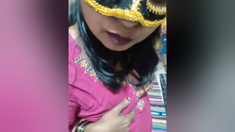 Media: A video of a woman with dark hair and a black, yellow-gold mask, wearing a pink sari with intricate embroidery, touching her chest, in a colorful indoor setting.