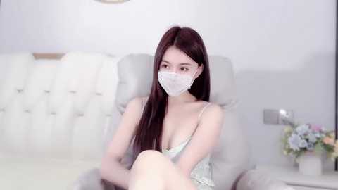 Media: A video of a young East Asian woman with long, straight dark hair, wearing a white face mask and a sheer, floral-patterned dress, sitting on a white, tufted sofa in a minimalist, softly lit room.