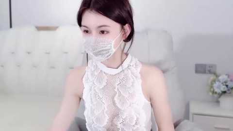 A video of an East Asian woman with fair skin, dark hair, and wearing a white lace sleeveless dress, face mask, and glasses, sitting on a white couch with a beige cushion, in a minimalist, modern living room.