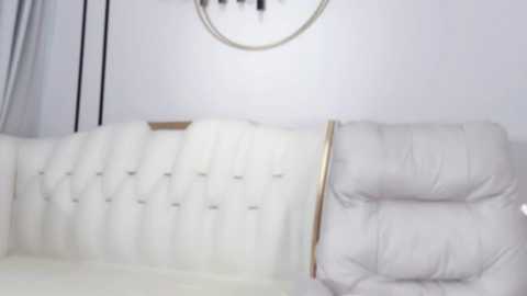 Media: A video of a modern, minimalist living room featuring a white, tufted leather sofa with a plush, cushioned armchair, against a white wall with a round, metallic light fixture.