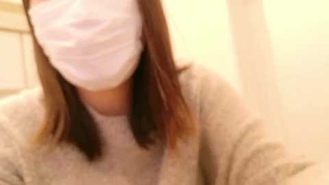 Media: A video of a person with light skin, wearing a white face mask, and a beige sweater, taken indoors with a soft, warm light.