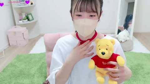 Media: Video of an Asian woman in a white mask, white shirt, and red collar, holding a Winnie the Pooh plush toy. Background shows a pink chair, green rug, and white shelves with toys.