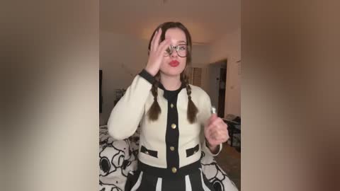 Media: Video of a young woman with fair skin and brown hair in pigtails, wearing glasses, a white cardigan with black trim, and a black skirt with a white pattern, standing in a dimly lit room.