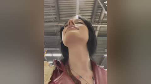 Media: Video of a woman with short black hair, wearing a maroon leather jacket, standing in a warehouse with a corrugated metal roof. She has her eyes closed and head tilted back, exuding a serene expression.