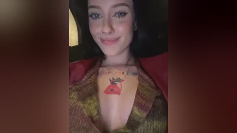 Media: Video of a smiling woman with light skin and dark hair, wearing a red top with a colorful, cartoonish tattoo of a red heart on her chest. Background is dimly lit, possibly indoors.