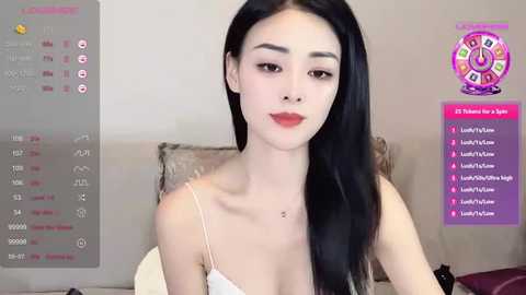 Media: Video of a slender, pale-skinned Asian woman with long black hair, wearing a white spaghetti strap top, sitting on a beige couch. Background includes a purple chat bubble screen and a digital clock.