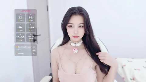 Media: Video of an East Asian woman with long black hair, wearing a pink lace top and silver choker with a heart pendant. She sits in a room with a white door and a digital thermometer on the wall.