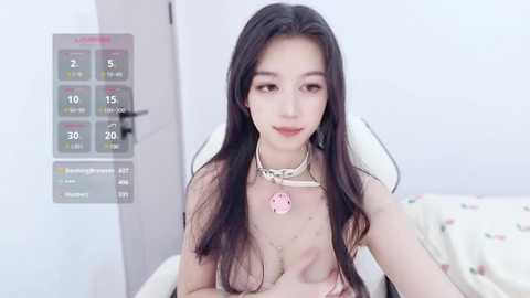 Media: A video of an East Asian woman with long, straight black hair, wearing a sheer, see-through top revealing her breasts, sitting on a bed with a white comforter. She has a pink pendant necklace and is in a bedroom with a digital calendar on the wall.