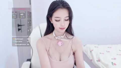 Media: Video of an East Asian woman with long black hair, fair skin, and a slender physique, wearing a pink choker with a heart pendant, sitting on a white gaming chair in a minimalist bedroom with a floral-patterned bedspread.