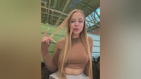 Video of a young blonde woman with long, straight hair, wearing a brown crop top and gray pants, holding a hairbrush in a modern, light-filled room with green walls and a metallic ceiling.