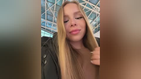 Video of a young, fair-skinned woman with long blonde hair, wearing a black jacket, posing indoors with geometric, modern architecture behind her.