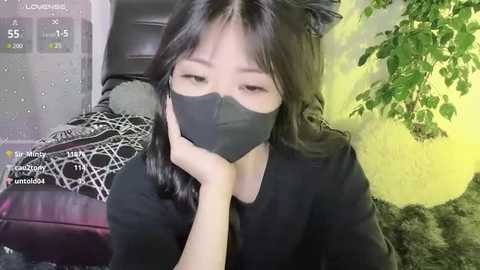 Video of an Asian woman with straight black hair, wearing a black mask and black shirt, resting her head on her hand, in a cozy, dimly-lit room with a green plant and black and white patterned blanket in the background.