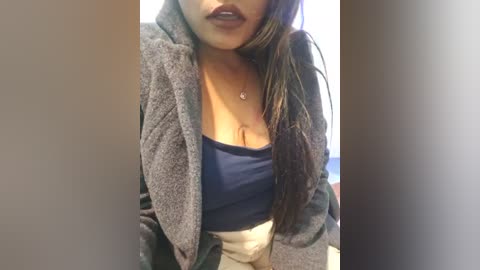 Media: Video of a young woman with long, straight dark hair, wearing a grey hoodie over a blue tank top, showcasing cleavage. She has a light brown skin tone, and her lips are painted dark.