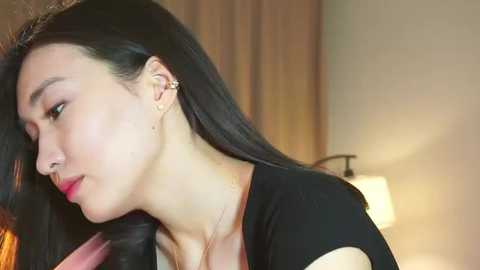 Media: Video of a woman with long black hair, fair skin, and a slender build, wearing a black top. She has a calm expression, looking downward. Background includes a beige wall and a lit lamp.