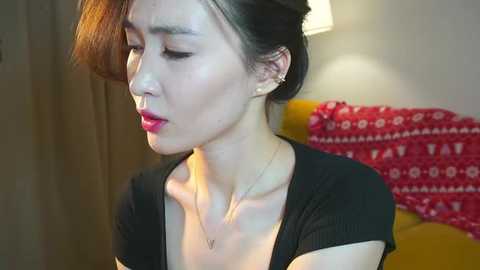 Video of a young Asian woman with fair skin, red lipstick, and dark hair styled in a bun. She wears a black top, and a pendant necklace. Background features a red pillow and a lamp.