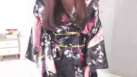 Media: Video of a woman in a black floral kimono with pink accents, wearing a yellow obi belt, standing in a minimalist room with a white wall and wooden dresser.