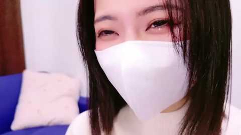 Media: Video of an Asian woman with straight black hair, wearing a white face mask, and a white top. Background shows a blue couch and a white pillow, hinting at a casual indoor setting.