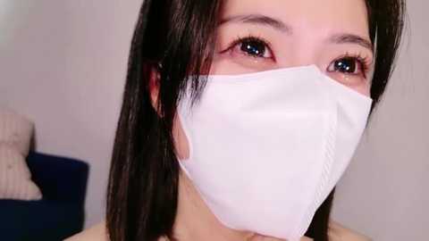 Media: Close-up video of an Asian woman with long black hair, wearing a white surgical mask, revealing her brown eyes and fair skin. Background features a blurred blue couch and beige wall.