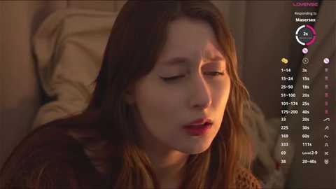 Video of a young woman with long brown hair, closed eyes, and open mouth, possibly crying, in a dimly lit room. Background shows a bed with beige pillows and a pink heart icon on a screen.