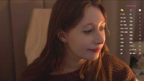 Video of a young Caucasian woman with light skin and straight, shoulder-length brown hair, wearing a brown knitted sweater. She has a serene expression, looking downward. Background includes a beige wall and a clock displaying 7:10.