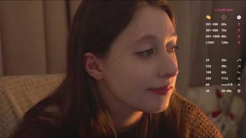 Video of a young girl with fair skin, brown hair, and a light brown sweater, looking contemplative in a cozy, dimly lit room.