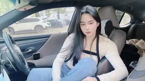 Media: Video of a young East Asian woman with long black hair, wearing a beige top and blue jeans, sitting in a car with a parked SUV visible outside the window.