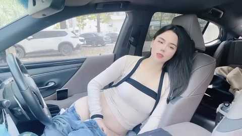 Media: Video of an attractive East Asian woman with long black hair, wearing a white crop top and blue jeans, reclining seductively in a car seat, with a dog sitting next to her.