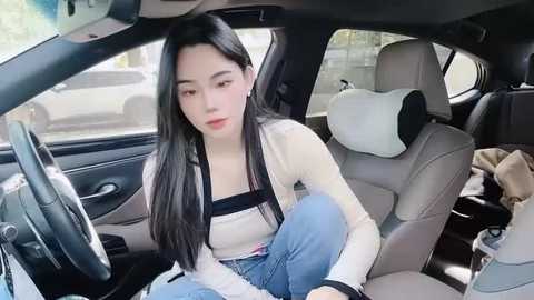 Media: Video of an East Asian woman with long black hair, fair skin, and a slender physique, wearing a white top and blue jeans, sitting in the driver's seat of a modern car.