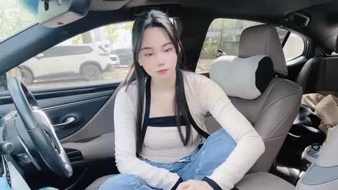 Media: Video of an East Asian woman with long black hair, wearing a white long-sleeve top and light blue jeans, sitting in a modern car with beige leather seats, looking introspective.