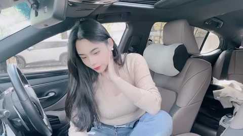 Media: Video of an Asian woman with long black hair, wearing a beige sweater and jeans, sitting in a modern car, adjusting her earring.