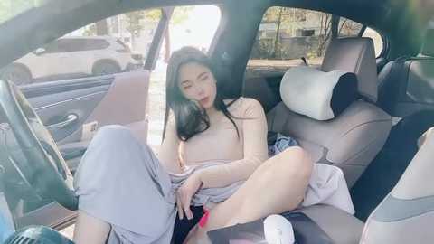 Media: Video of an Asian woman with long black hair, wearing a beige top, grey pants, and white shoes, reclining seductively in a car seat, legs spread, masturbating.