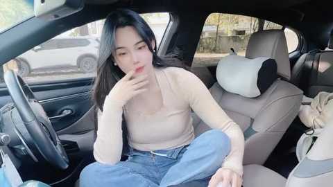 Media: Video of an Asian woman with long black hair, wearing a beige long-sleeve top and blue jeans, sitting in a car seat, hand to her mouth, looking contemplative.