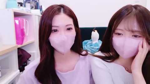 Media: Video of two young Asian women with long brown hair wearing face masks, sitting closely together. Background shows a white shelf with pink and blue objects.