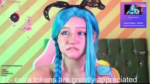 Media: Video of a young woman with bright blue hair, wearing a black lace headband, in a colorful, futuristic room with a green couch. Text overlay: \"22 - extra tokens are greatly appreciated.\