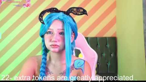 Video of a pale-skinned woman with bright blue hair in twin braids, wearing bunny ears and a lace choker. She has a tattoo on her right shoulder. Background features a pink and yellow striped wall. Text overlay: \"22 - extra tokens are greatly appreciated\".