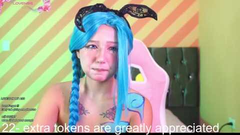 Media: Video of a young woman with vibrant blue hair and bunny ears, wearing a strapless top. The background features a striped wall and a pink gaming chair. Text overlays the image, \"22 extra tokens are greatly appreciated.\