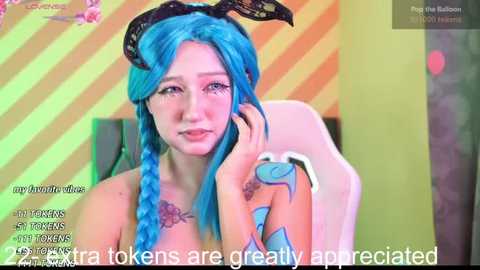 Media: Video of a young woman with vibrant blue hair, wearing black bunny ears, sitting in a gaming chair. She has a tattoo on her shoulder. The background is a colorful, striped wall. Text overlays her image.