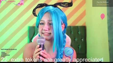 Media: Video of a smiling young woman with blue hair, wearing bunny ears, holding a microphone, against a yellow-striped wall background. Text: \"22 extra tokens are greatly appreciated.\