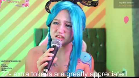 Media: Video of a woman with bright blue hair and black bunny ears singing into a microphone. She's seated on a green chair, with a yellow and orange striped background. Text at the bottom reads, \"22 extra tokens are greatly appreciated.\