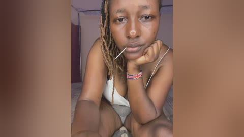 Media: Video of a young Black woman with medium brown skin, wearing a white tank top, seated on a bed in a dimly lit room. She has long, thin dreadlocks, a beaded bracelet, and rests her chin on her hand, looking contemplative.