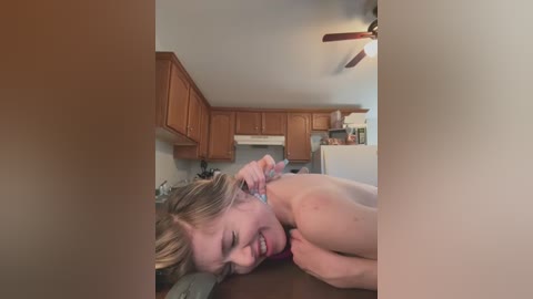 Media: Video of a nude blonde woman lying on a kitchen counter, smiling, in a small, dimly lit kitchen with wooden cabinets and a ceiling fan.