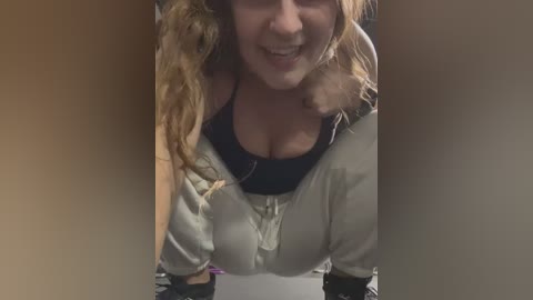 Media: A video of a smiling woman with curly blonde hair, wearing a beige hoodie over a black top, partially obstructed by two hands.