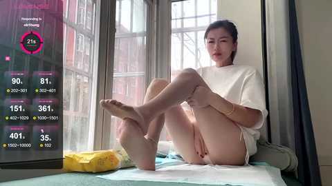 Video of an Asian woman in a white t-shirt and panties, sitting on a bed by a window, displaying health stats on a screen.