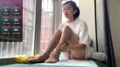 Video of an Asian woman in a white t-shirt and shorts, sitting by a window, with a digital overlay displaying a fitness app.