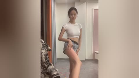 Media: A video of a slender, light-skinned woman with long dark hair, wearing a white crop top and denim shorts, standing in a hallway. Background includes a door, wall, and a colorful rug.