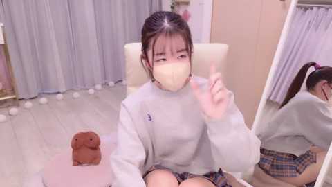 Media: Video of an East Asian woman with long black hair in a ponytail, wearing a gray sweatshirt and plaid skirt, sitting on a plush pink chair in a soft-lit room with beige walls, a mirror, and a teddy bear.