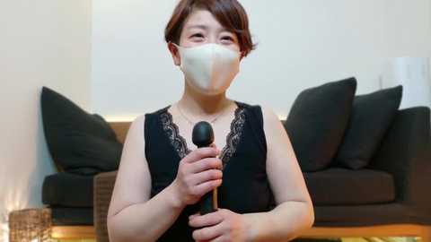 Media: Video of an East Asian woman in a black lace-trimmed top, wearing a face mask, holding a microphone in a living room with a dark couch and white blinds.