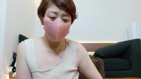 Media: Video of an East Asian woman with short brown hair, wearing a beige tank top, and pink face mask, sitting indoors, with a black couch and beige wall in the background.